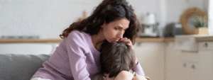 A woman with wavy hair, wearing a lavender sweater, comforts a child on her lap in a bright kitchen, conveying care and support.