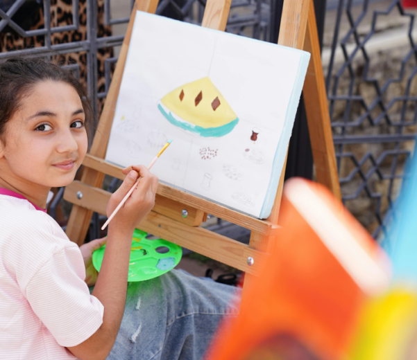 A young girl sits outdoors, holding a paintbrush and palette, working on a painting of a yellow object on an easel, and looking back at the camera.
