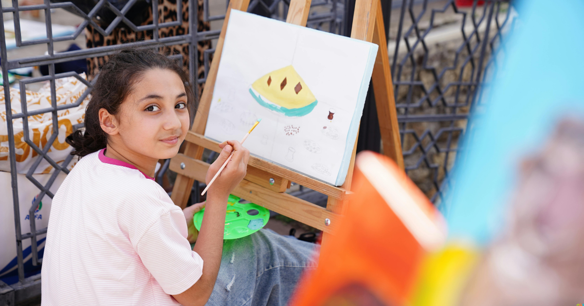 A young girl sits outdoors, holding a paintbrush and palette, working on a painting of a yellow object on an easel, and looking back at the camera.
