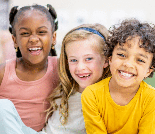 Three children sit closely, smiling joyfully. The girl on the left wears a pink dress, the middle one has a headband, and the boy wears a yellow shirt.