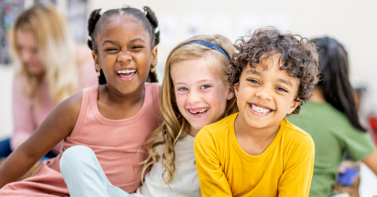 Three children sit closely, smiling joyfully. The girl on the left wears a pink dress, the middle one has a headband, and the boy wears a yellow shirt.