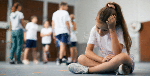 A sad, young girl with a ponytail sits on a gym floor, her head resting on her hand and her gaze downcast. She is wearing a white T-shirt and shorts. In the blurry background, other children are standing and moving around, suggesting a gym class or sports activity.