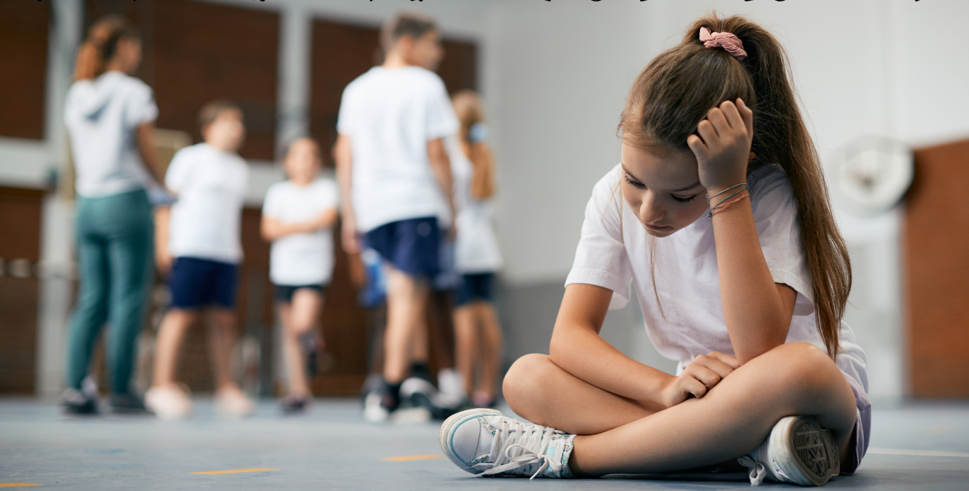 A sad, young girl with a ponytail sits on a gym floor, her head resting on her hand and her gaze downcast. She is wearing a white T-shirt and shorts. In the blurry background, other children are standing and moving around, suggesting a gym class or sports activity.