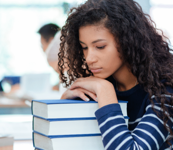 A young african american woman with curly hair rests her head on a stack of books in a library. She appears sad, wearing a striped shirt. Soft lighting and a blurred background suggest a quiet study environment.