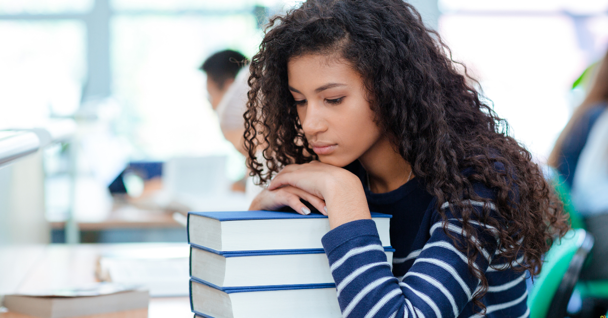A young african american woman with curly hair rests her head on a stack of books in a library. She appears sad, wearing a striped shirt. Soft lighting and a blurred background suggest a quiet study environment.