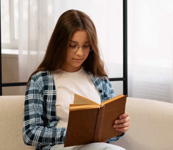 A young girl with glasses and a plaid shirt reads a book on a cozy beige bean bag, near a bright window with sheer curtains. She appears focused and relaxed.