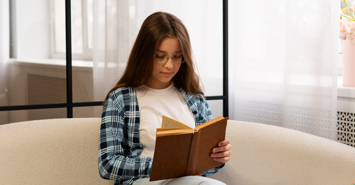 A young girl with glasses and a plaid shirt reads a book on a cozy beige bean bag, near a bright window with sheer curtains. She appears focused and relaxed.