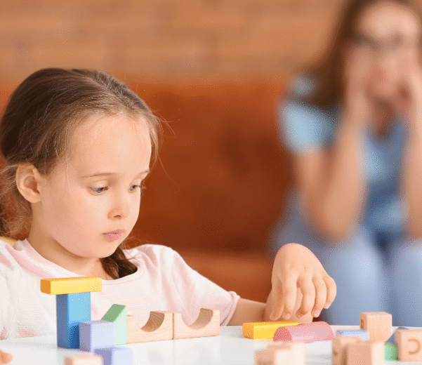 A young girl sits at a table playing with wooden blocks while an adult woman sits on a couch in the background, watching her.