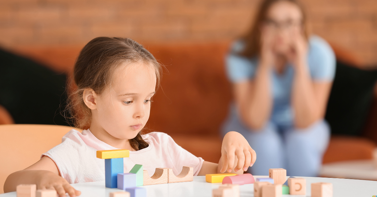 A young girl sits at a table playing with wooden blocks while an adult woman sits on a couch in the background, watching her.