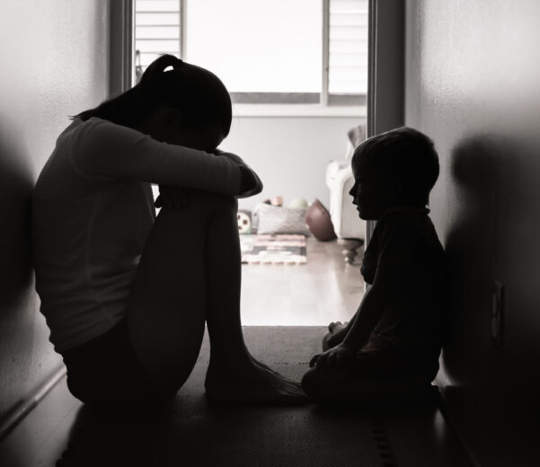 Silhouetted in a dim hallway, a child with head on knees sits facing a smaller child. Light from a doorway reveals toys in the background. The mood is somber.