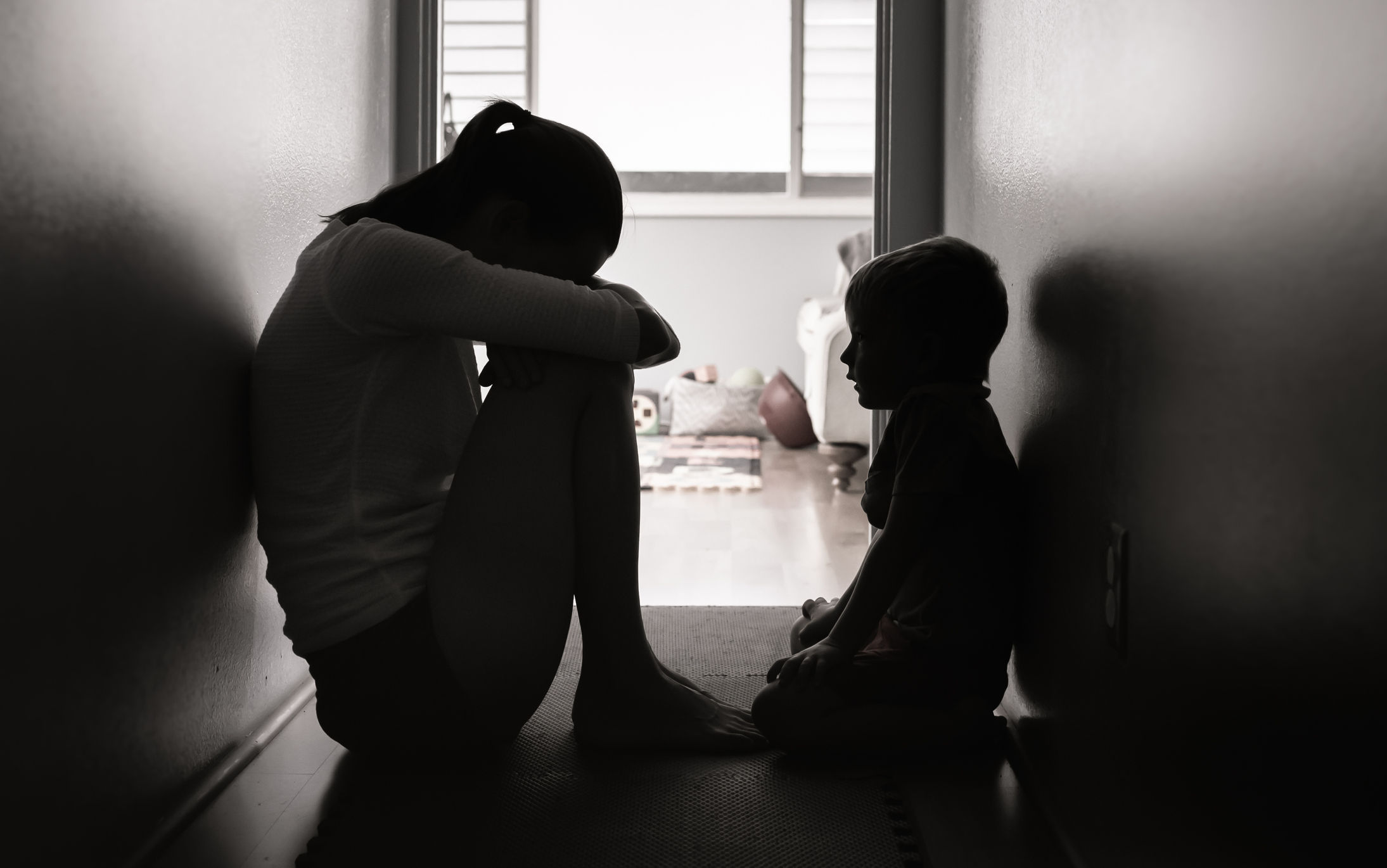 Silhouetted in a dim hallway, a child with head on knees sits facing a smaller child. Light from a doorway reveals toys in the background. The mood is somber.